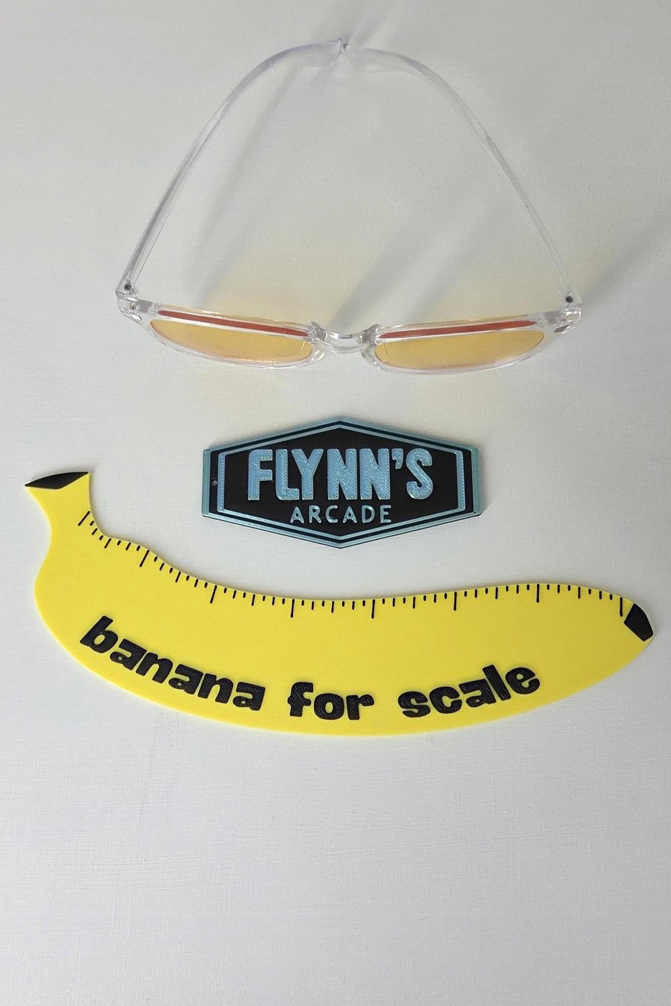 Clear sunglasses with yellow lenses on a white background, featuring a 'Flynn's Arcade' logo and a banana-shaped ruler.